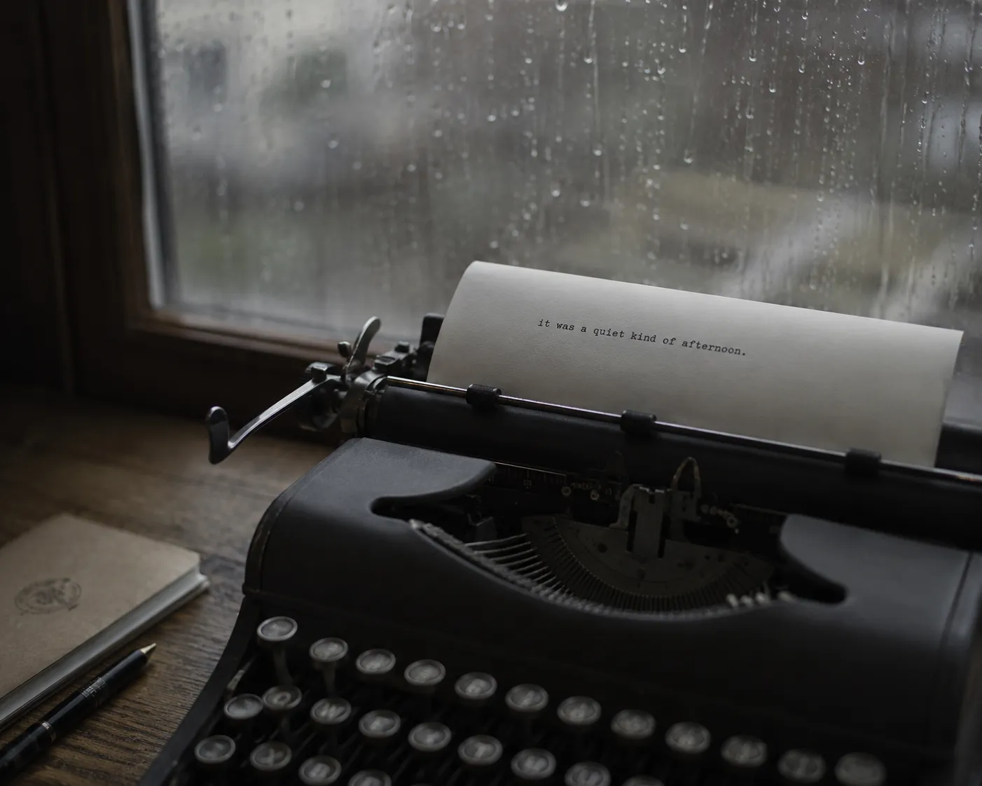Une machine a ecrire ancienne pres d'une fenetre couverte de pluie, une feuille engagee avec une seule ligne tapee, lumiere grise et douce.