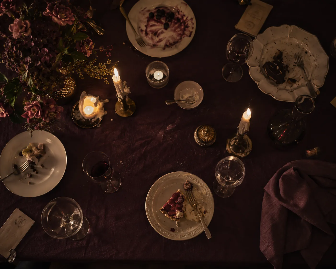 Vue de dessus d'une table de diner en fin de soiree, nappe prune, assiettes de dessert, verres a moitie pleins et bougies presque fondues.